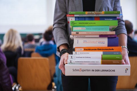 Bild, auf dem eine Person mit einem Stapel Büchern in den Händen in einem Hörsaal steht. Im Hintergrund sind verschwommen eine Tafel und Stühle zu sehen, auf denen mehrere Personen sitzen.