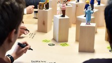 Personen schreiben mit Marker auf großer Fläche, auf der Holzfiguren (Bürger der Demokratie) stehen