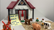 Mit Spielfiguren wurde die folgende Film-Szene aufgebaut: Kinder und Bauernhoftiere stehen im Heu vor einem Schulgebäude, an dem steht "nach der Schule: Arbeit auf dem Hof". Links davon sind ein Fußball und andere Hobby-Utensilien mit einem roten Kreuz bi