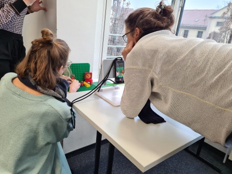 Drei Personen betrachten ihre Video-Szene, die mit Spiel-Figuren auf einem Tisch aufgebaut wurde. Sie blicken durch eine Smartphone-Kamera auf die aufgebaute Szene.