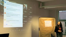 In der rechten Bildhälfte steht die Dozentin Melanie Wohlfahrt, die auf eine Beamer-Projektionsfläche im linken Bildbereich blickt. Hier kann man das Feedback mehrerer Teilnehmender lesen, die sich im Zoom-Chat zur Werkstattwoche wohlwollend äußern.