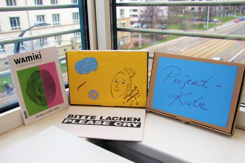 Man sieht zwei verschiedene bunte Lernkisten, die auf einem Tisch stehen und vor ein Fenster an der DIU gelehnt sind, sodass man im Hintergrund den Blick nach draußen bekommt.