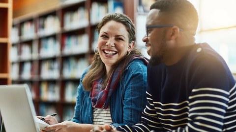 Foto von zwei lachenden Studenten, die in der Bibliothek zusammen am Laptop sitzen und lernen.