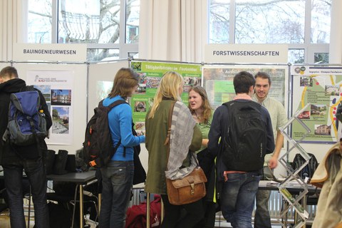 Das Foto zeigt einen Raum mit einigen Studierenden. Sie stehen vor Tischen und Informationstafeln zu verschiedenen Studiengängen. Studierende dieser Studiengänge stehen vor den Tafeln und erklären den interessierten Schüler:innen etwas. Man erkennt die Aufschrift "Bauingenieurwesen" und "Forstwissenschaften" über den Tafeln.