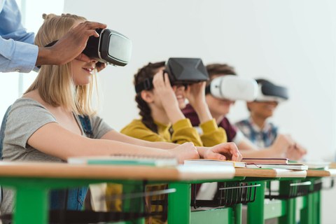 Das Foto zeigt vier Jugendliche. Sie sitzen an Tischen in einem Klassenzimmer und tragen eine VR-Brille.
